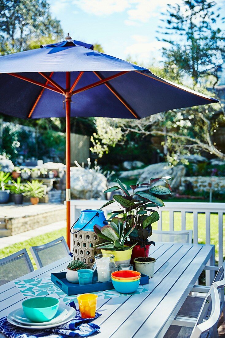 Blue parasol above terrace table set with colourful bowls; sunny garden in background