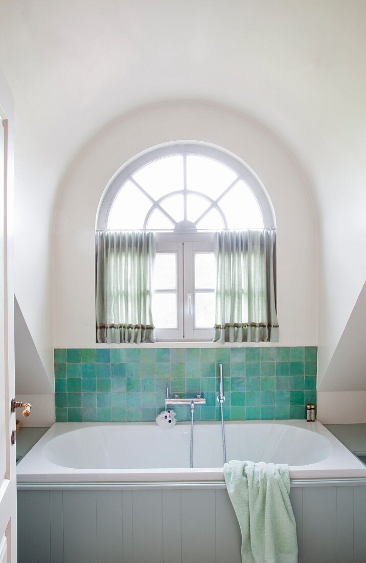 Bathtub against partially tiled wall in shades of green in arched dormer window with semicircular transom window