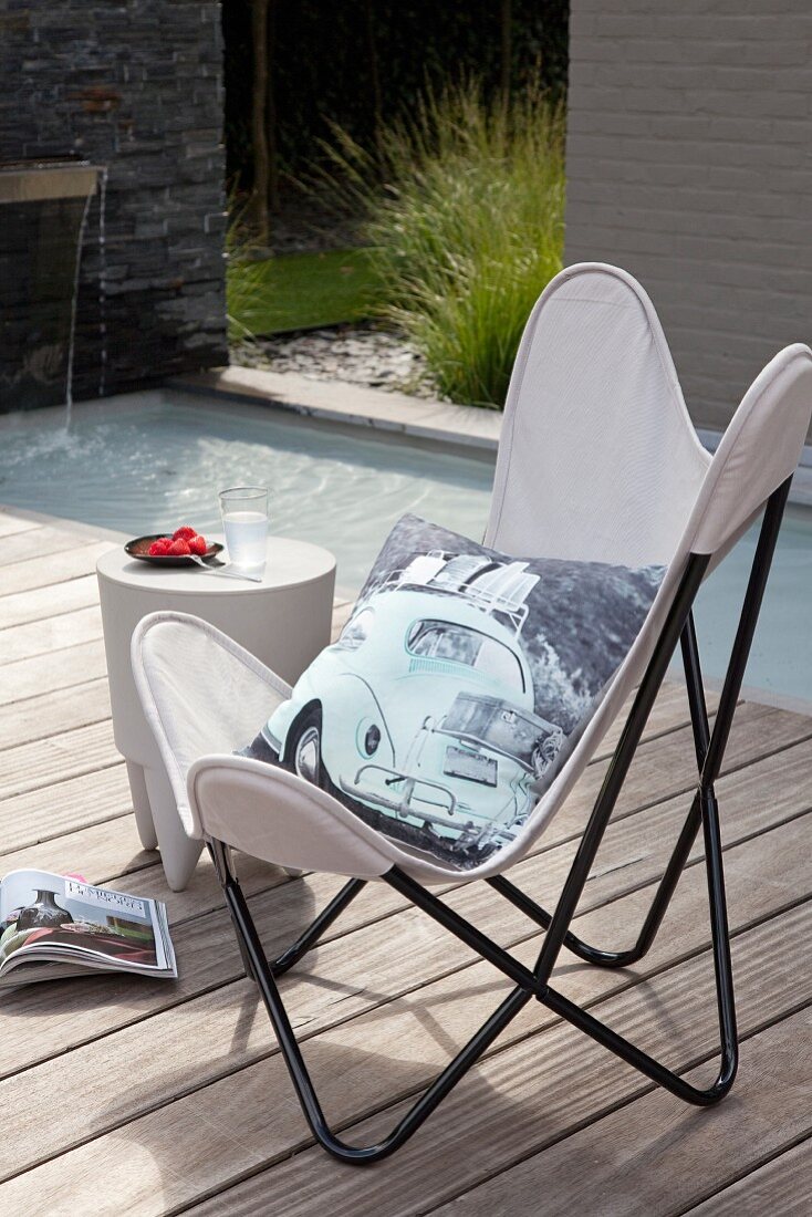 Printed cushion with car motif on white Butterfly chair and stool used as side table on sunny wooden deck next to pool