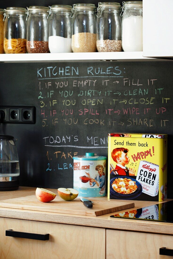 Storage jars on white kitchen shelf above writing on chalkboard splashback and retro tins