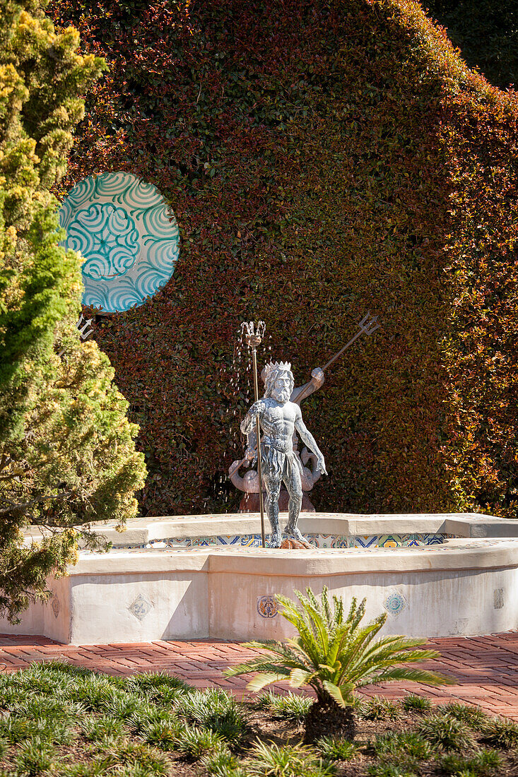 Sculpture of Neptune in pool in front of painted dish hung on clipped hedge
