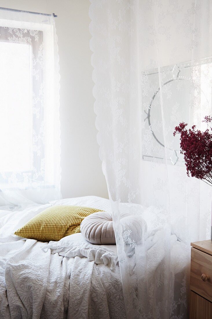 Romantische, weiße Spitzengardine vor Bett mit Tagesdecke und Kissen