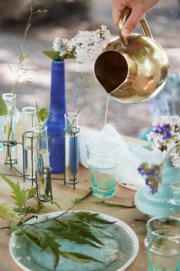 Pouring water from brass jug into glass on table set with summer flowers