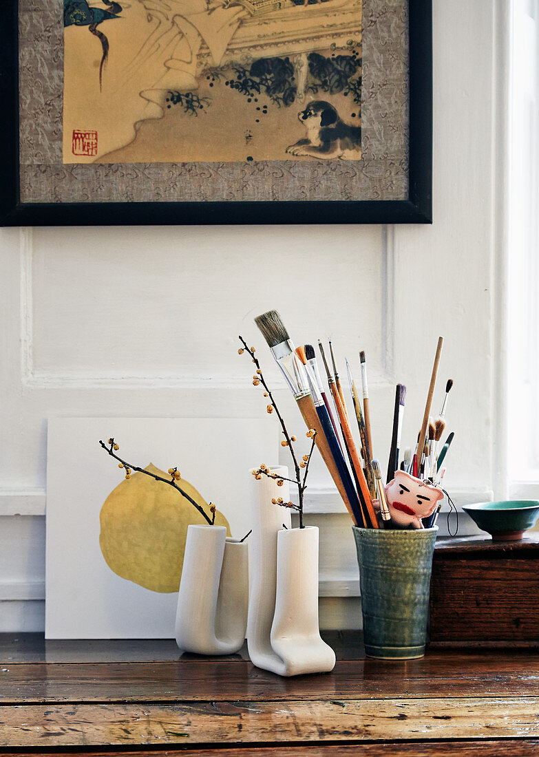 Pinselsammlung in Keramikbecher neben künstlerischen, weißen Vasen mit getrockneten Zweigen