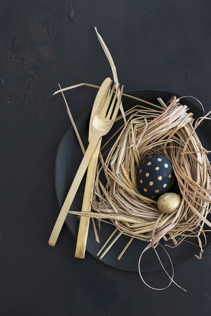 Oster-Tischdekoration auf schwarzem Teller mit Goldbesteck, Nest und schwarz-gold bemalten Eiern