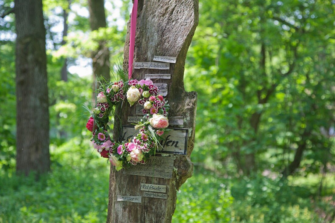Blumenkranz hängt an einem Brett mit kleinen Schildern im Wald