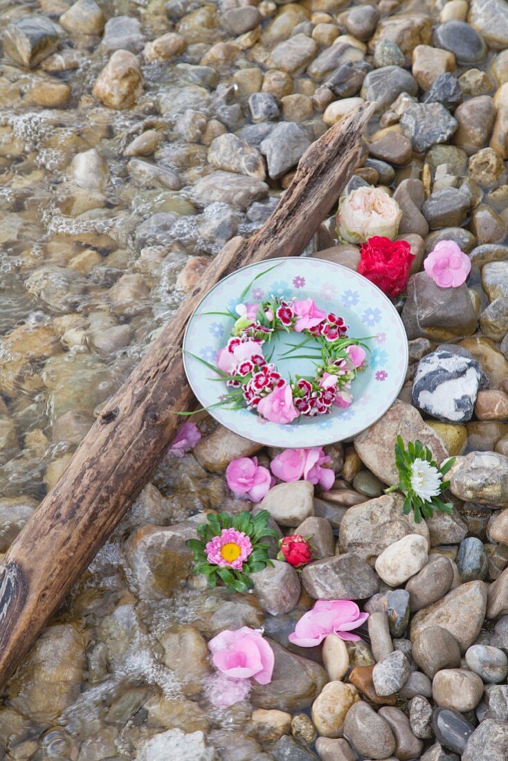 Teller mit Blumenkranz und Blüten im Kiesbett am Wasser