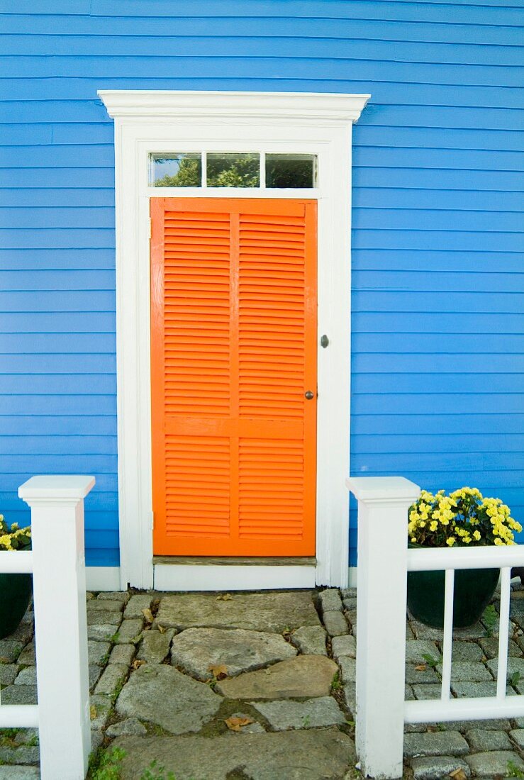 Orangene Lamellentür an blauem Holzhaus mit gepflastertem Eingang
