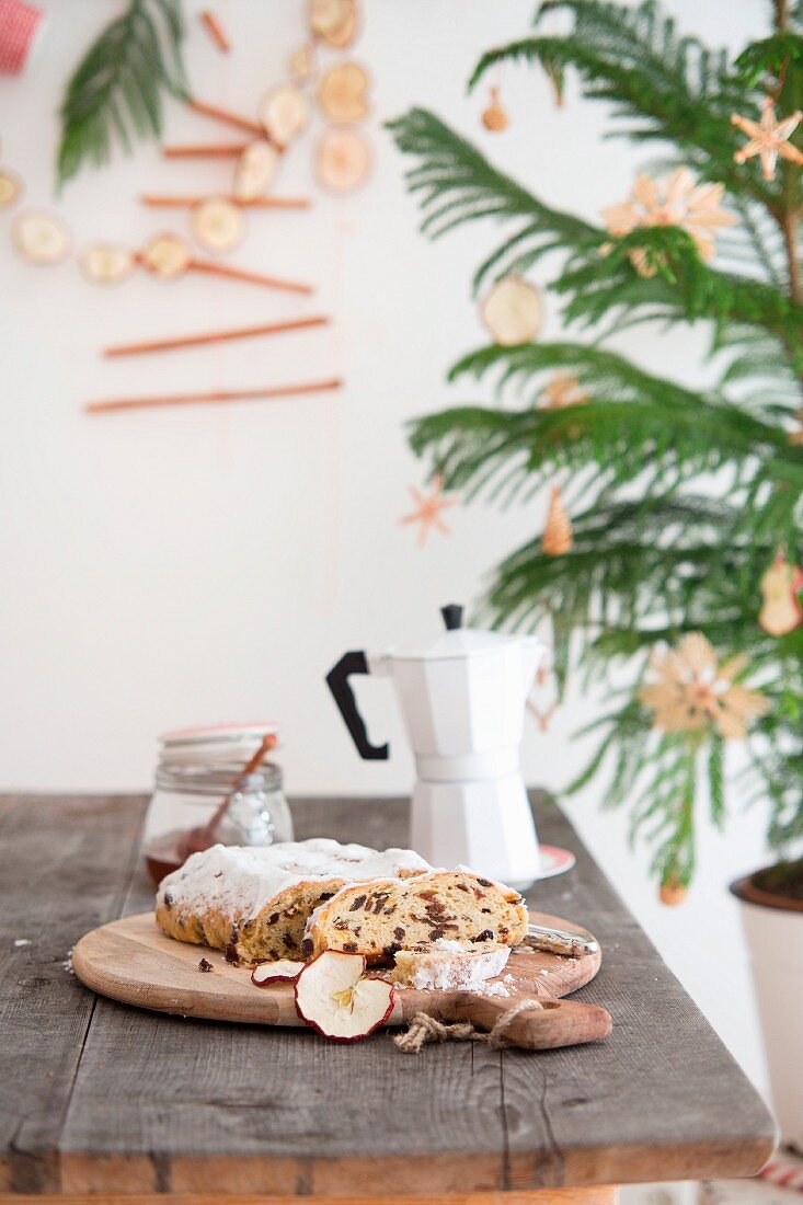 Christstollen, Honigglas und Espressokanne auf Holztisch, daneben Tannenbäumchen mit Strohdeko