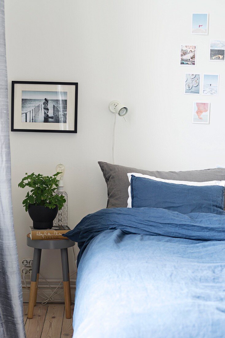 Schlafzimmer mit blauer Bettwäsche, Hocker als Nachttisch und verschiedenen Fotos als Wanddekoration