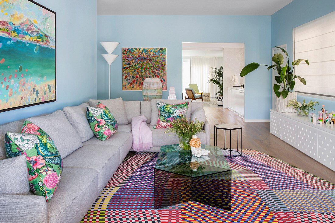 Wohnzimmer mit pastellblauen Wänden, grauer Eckcouch und Glas-Couchtisch auf farbenfroh gemustertem Teppich