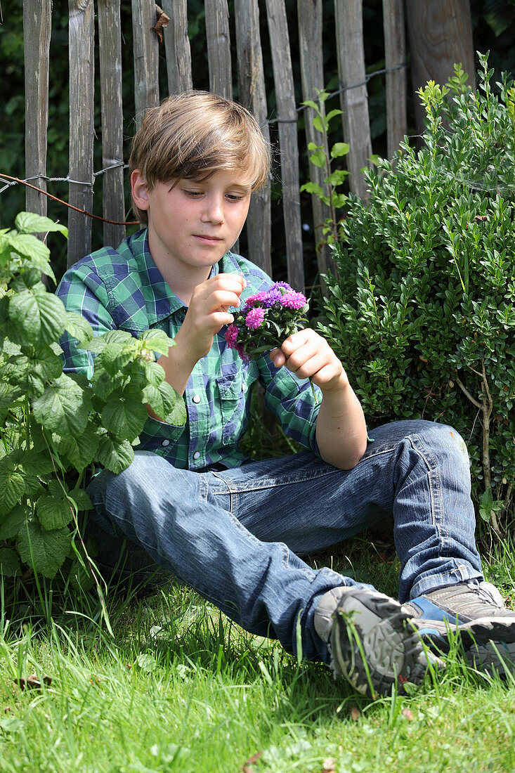 Boy holding posy of asters sitting on grass next to fence