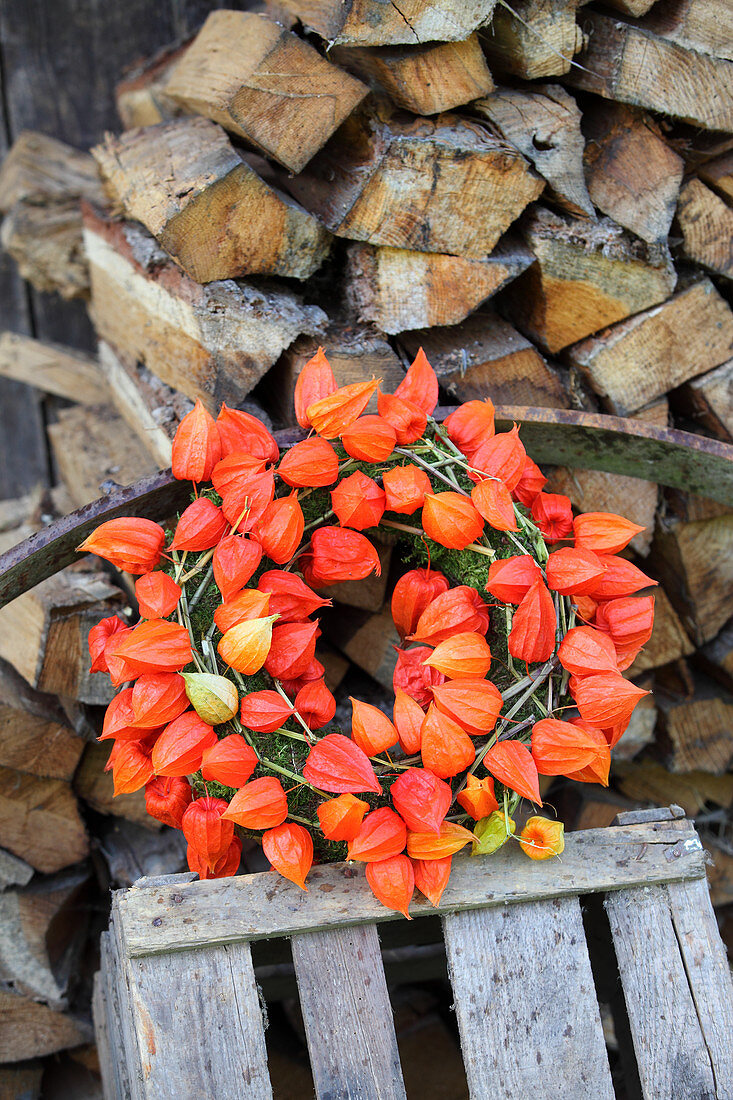 Mooskranz mit Lampionblumen auf einer Kiste vor dem Holzstapel