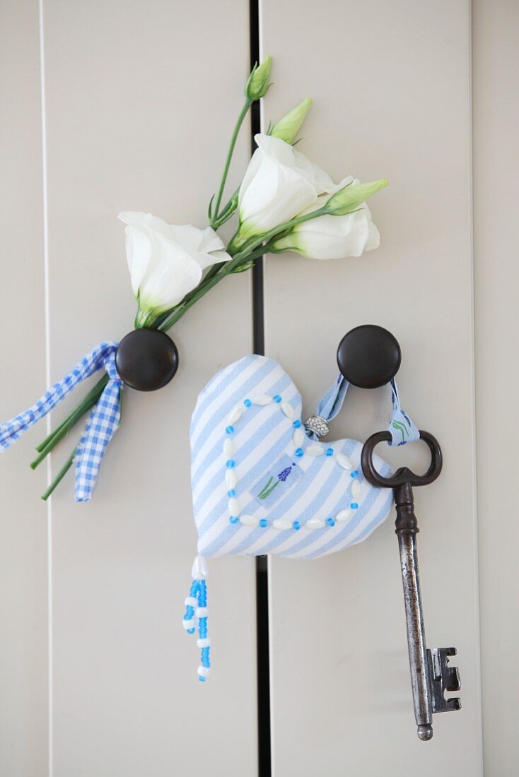 Pale blue, ornate, heart-shaped pendant and key next to white artificial flowers on cabinet door knobs