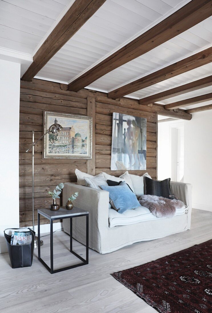 Helle Couch mit verschiedenen Kissen und Schaffell vor Holzwand mit Gemälden