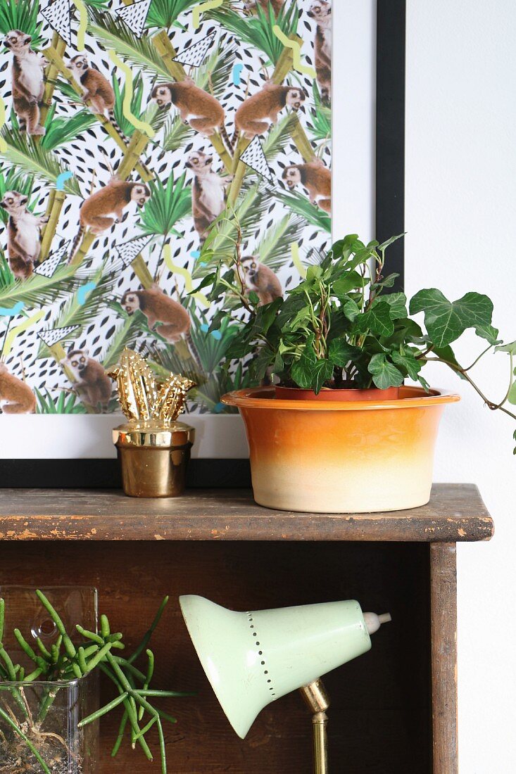Still-life arrangement of ivy next to gold cactus ornament in front of artwork with monkey motif