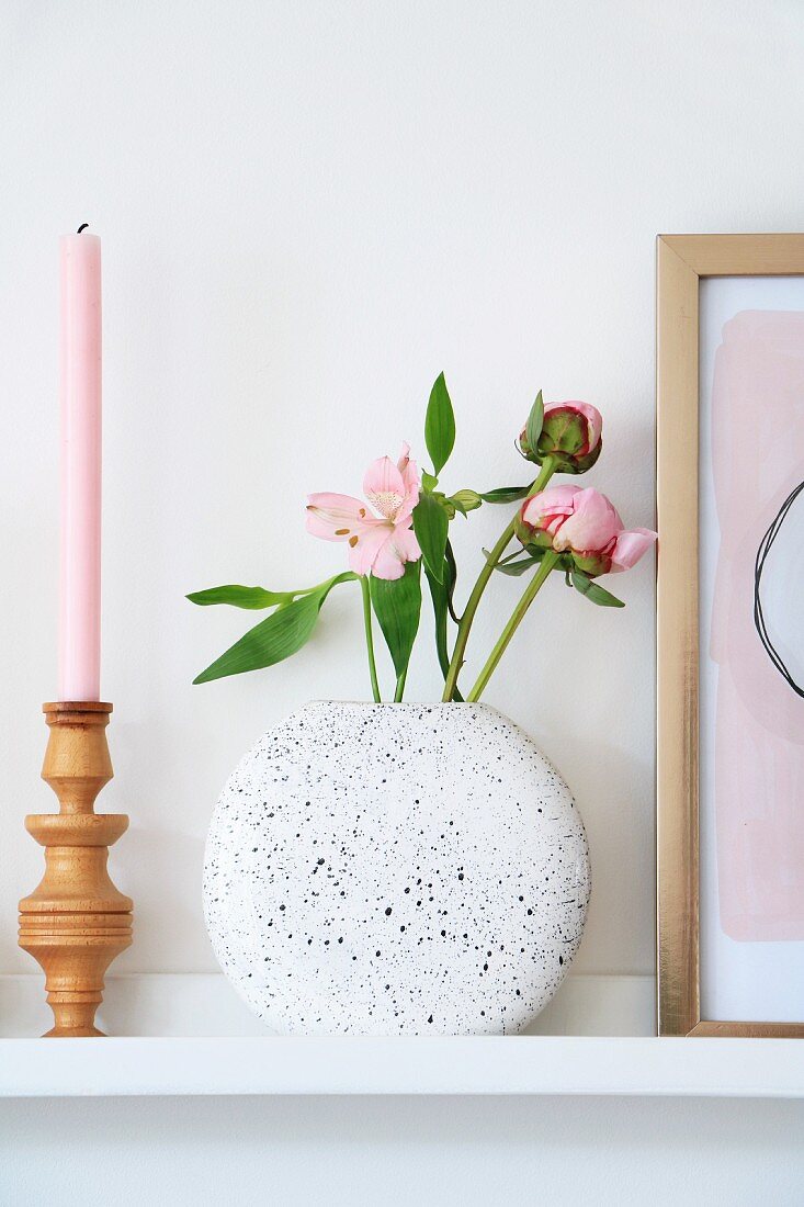 Gedrechselter Kerzenständer, rosa Kerze und gesprenkelter Vase mit Blumen auf Wandboard