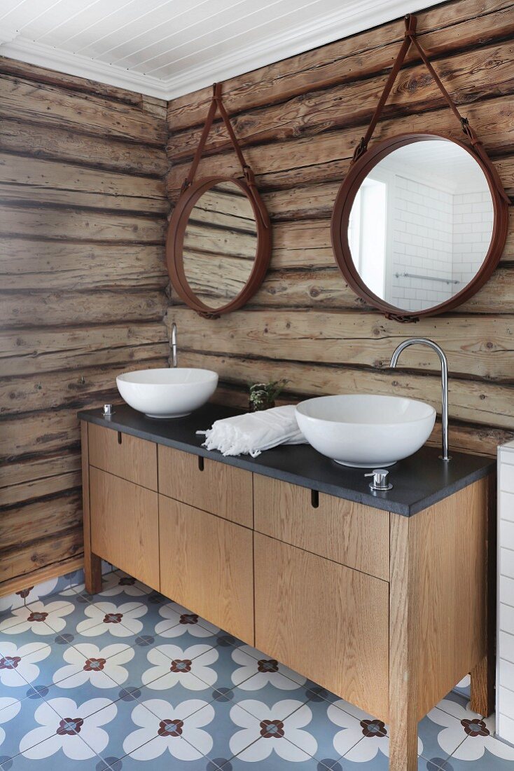Custom washstand with twin white washbasins against rustic log-cabin wall and cement floor tiles with floral pattern
