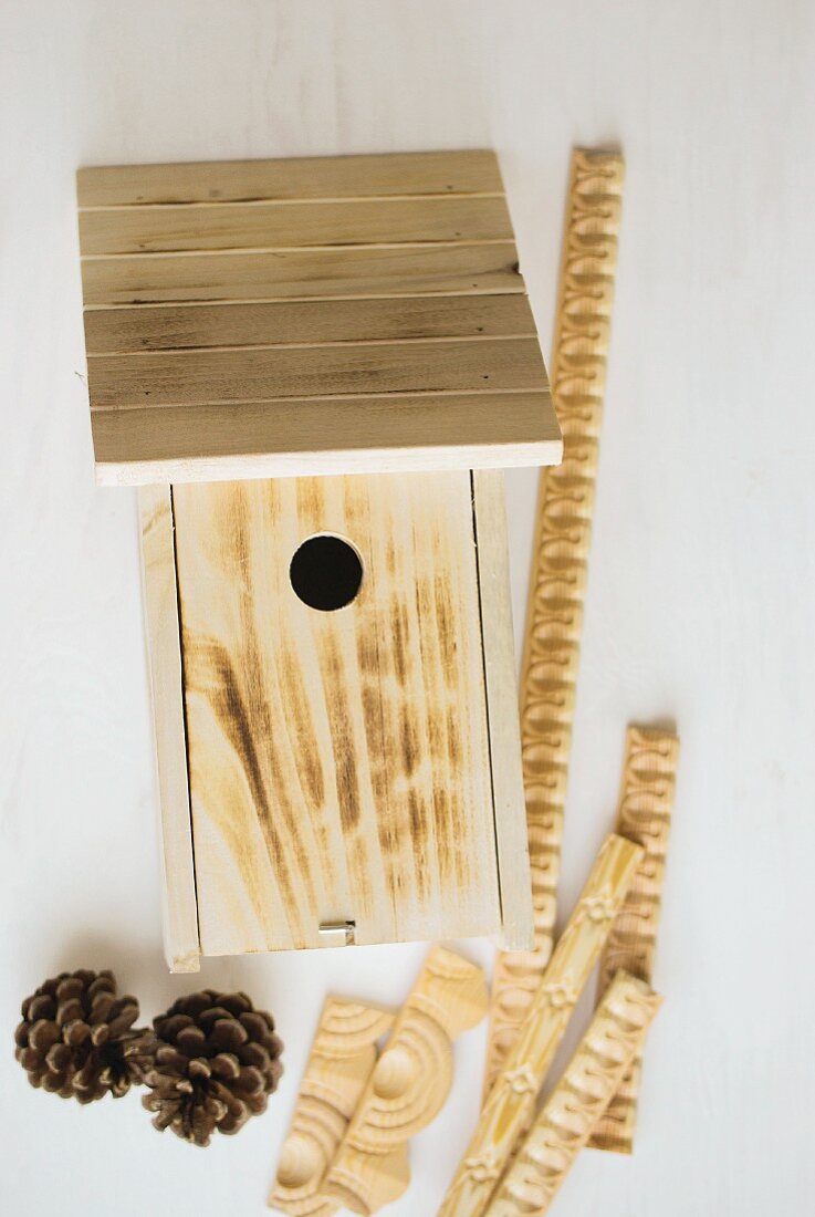 Traditional bird nesting box decorated with ornamental trim and pine cones on white surface