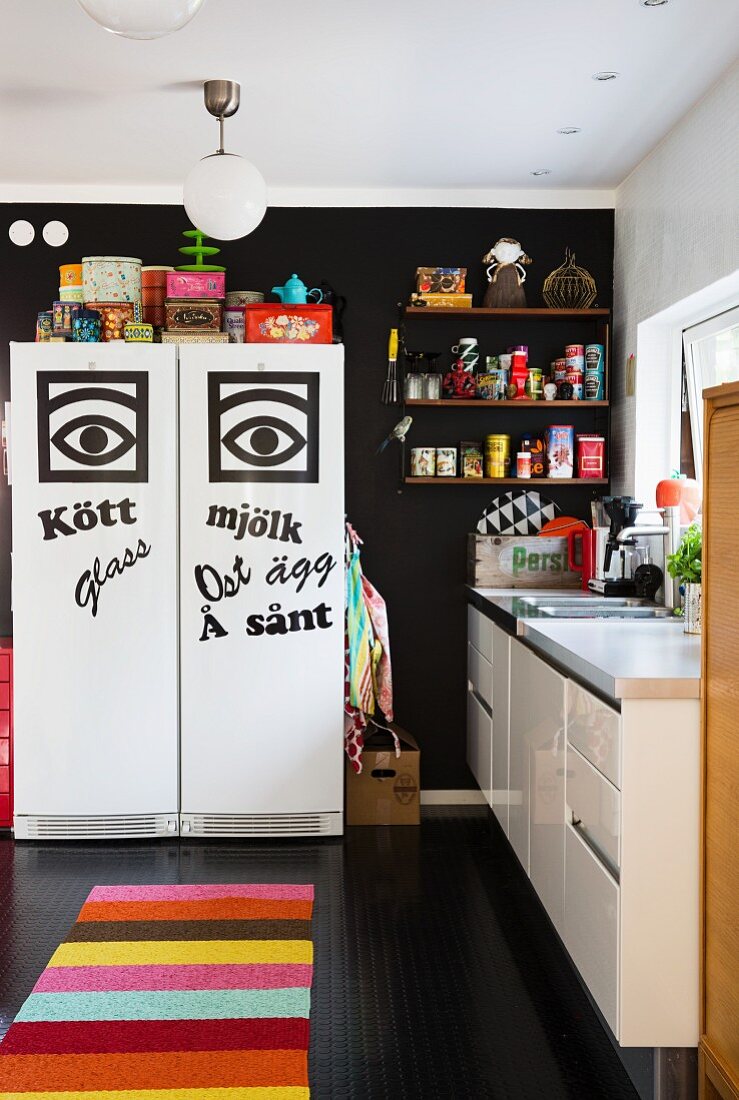 Kitchen with graphic decorations on fridge-freezer against black wall and colourful striped rug on black floor