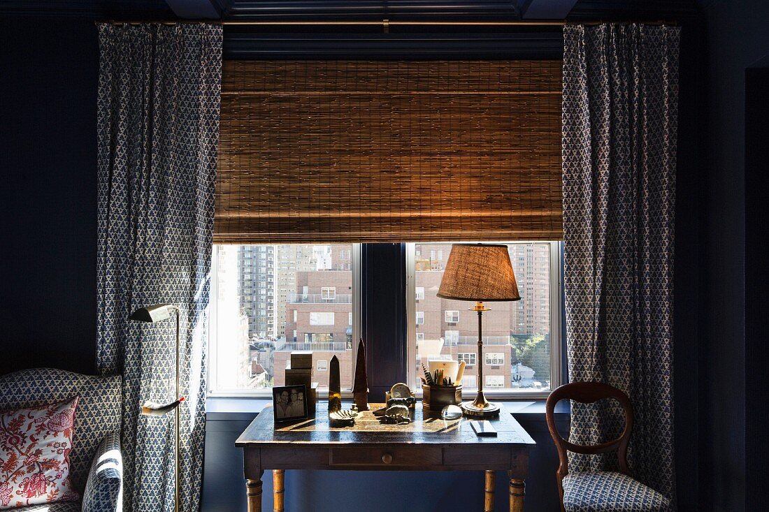Table lamp on antique desk in front of window with bamboo roller blind and matching blue and white patterned curtains, armchair and chair seat