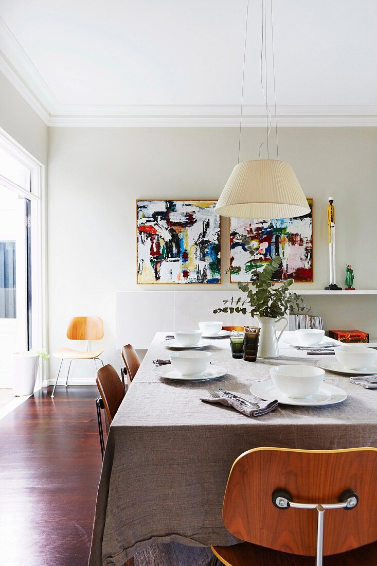 Klassiker Holzstühle um gedeckten Tisch in hellem Wohnraum mit Pendelleuchte und modernen Kunstgemälden
