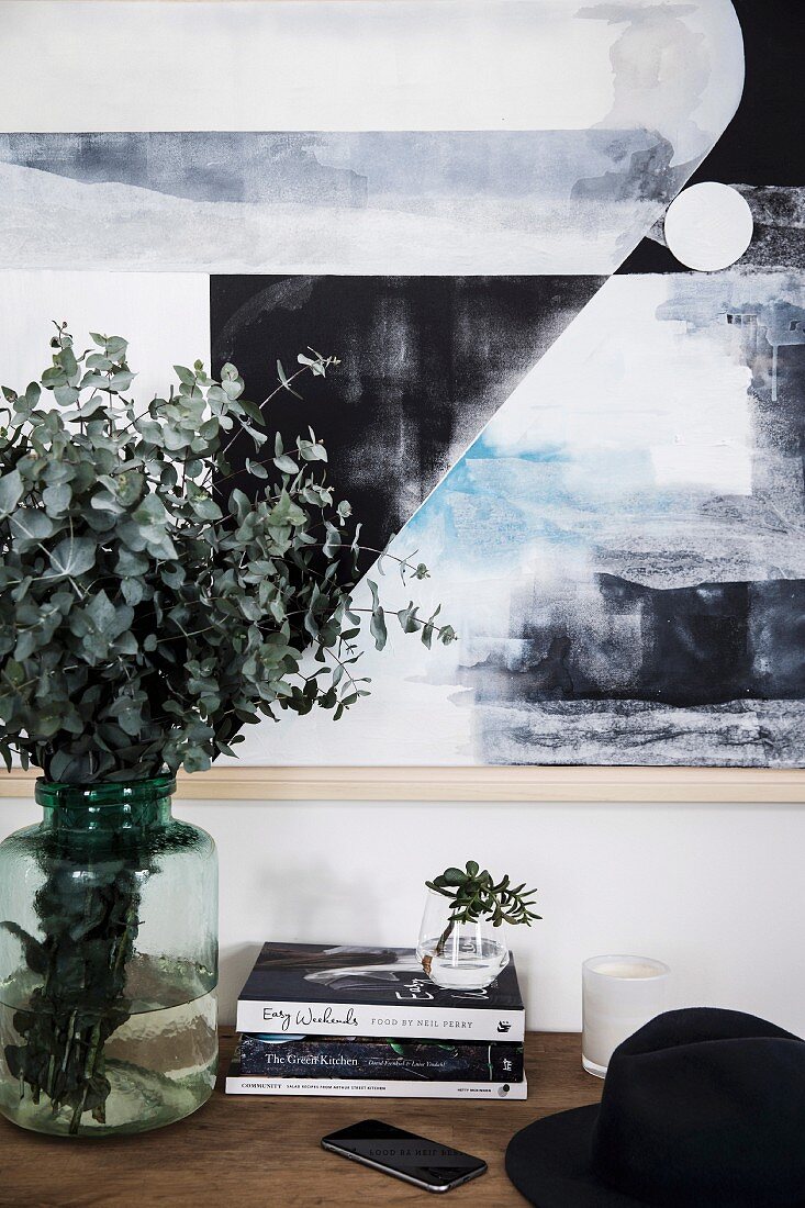 Blätterzweige in Glasvase, Bücherstapel und schwarzer Hut vor Wand mit modernem Bild