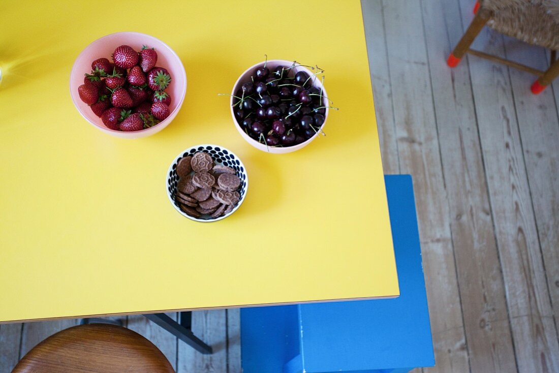 Drei Schälchen mit Erdbeeren, Kirschen und Schokokeksen auf gelber Tischplatte, darunter blauer Tritthocker