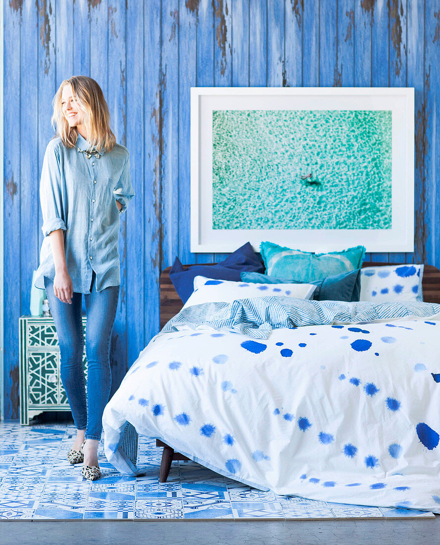 Doppelbett mit gemusterter Bettwäsche und blonde Frau vor blauer Stellwand