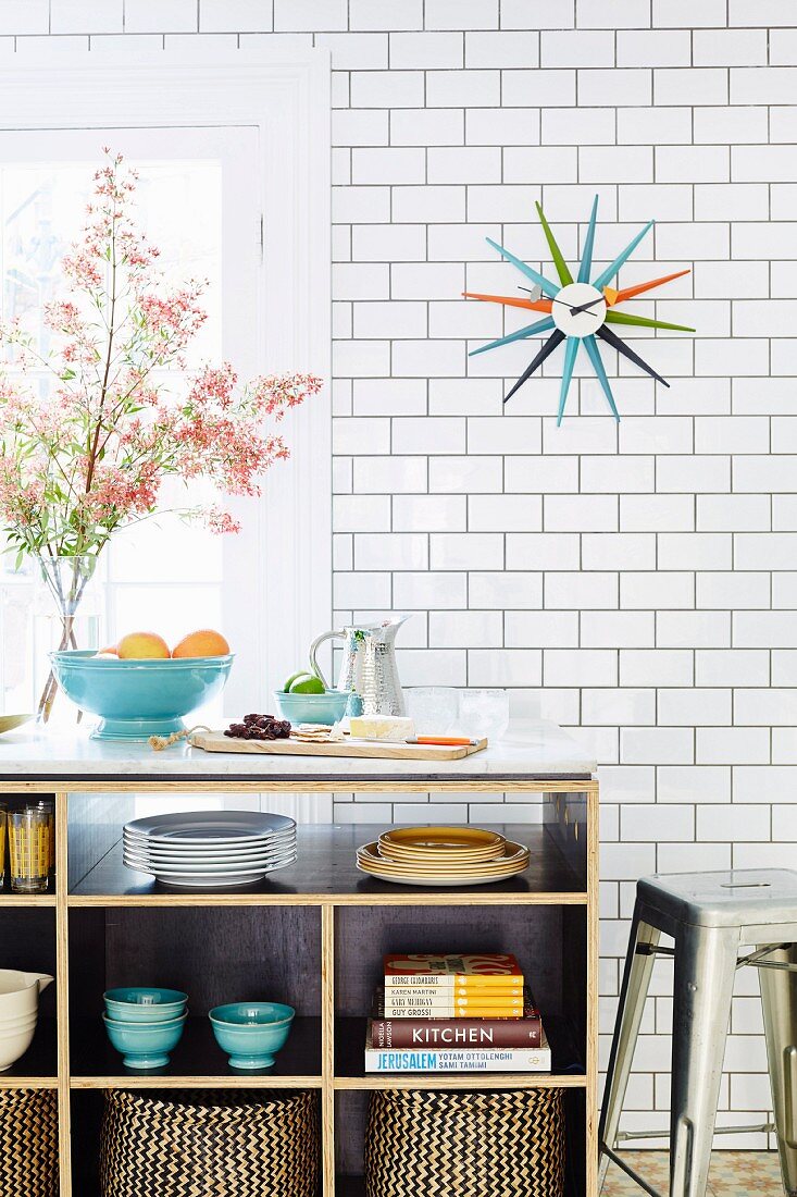 Shelf with dishes in front of white tiled wall, above wall clock with colored rays