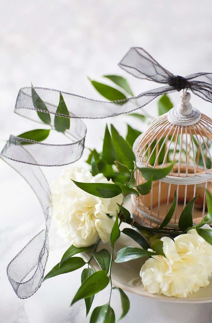 Black transparent ribbon draped around white carnations, leafy branches and ornamental birdcage