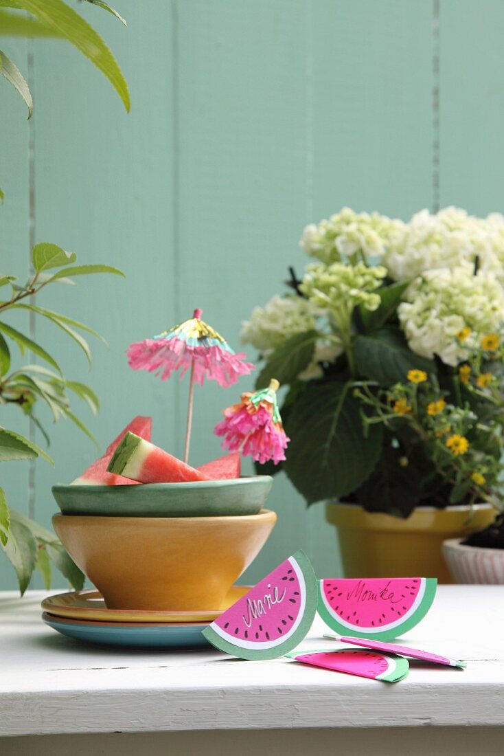 DIY-Tischkärtchen aus gefaltetem Papier in Melonenschnitzform auf Gartentisch neben Keramikschalen