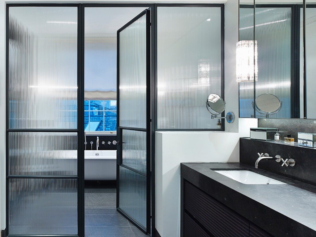 Steinverkleidetes Waschbecken mit Wandarmaturen und schwarzer Stahlrahmenglastür zum Badbereich