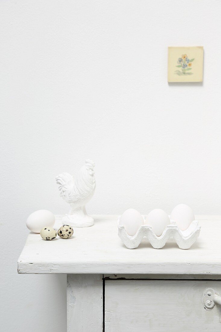 weiße Eier im Eierkarton, Wachteleier und Hahnfigur auf Kommode