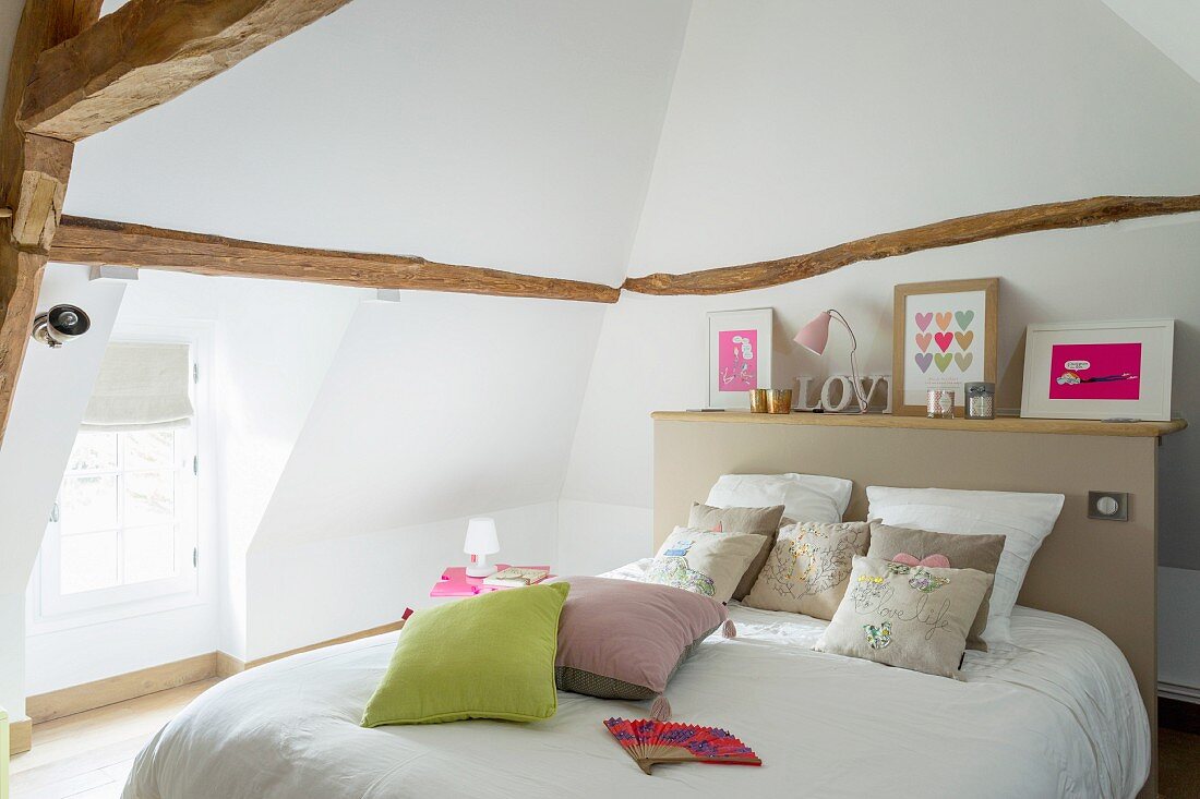 Double bed with shelf on top of beige headboard in converted attic with dormer window
