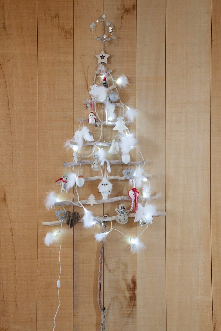 Selbstgebastelter Weihnachtsbaum aus dünnen Ästen, dekoriert mit Lichterkette, Federn und Baumanhängern, an Holzwand aufgehängt