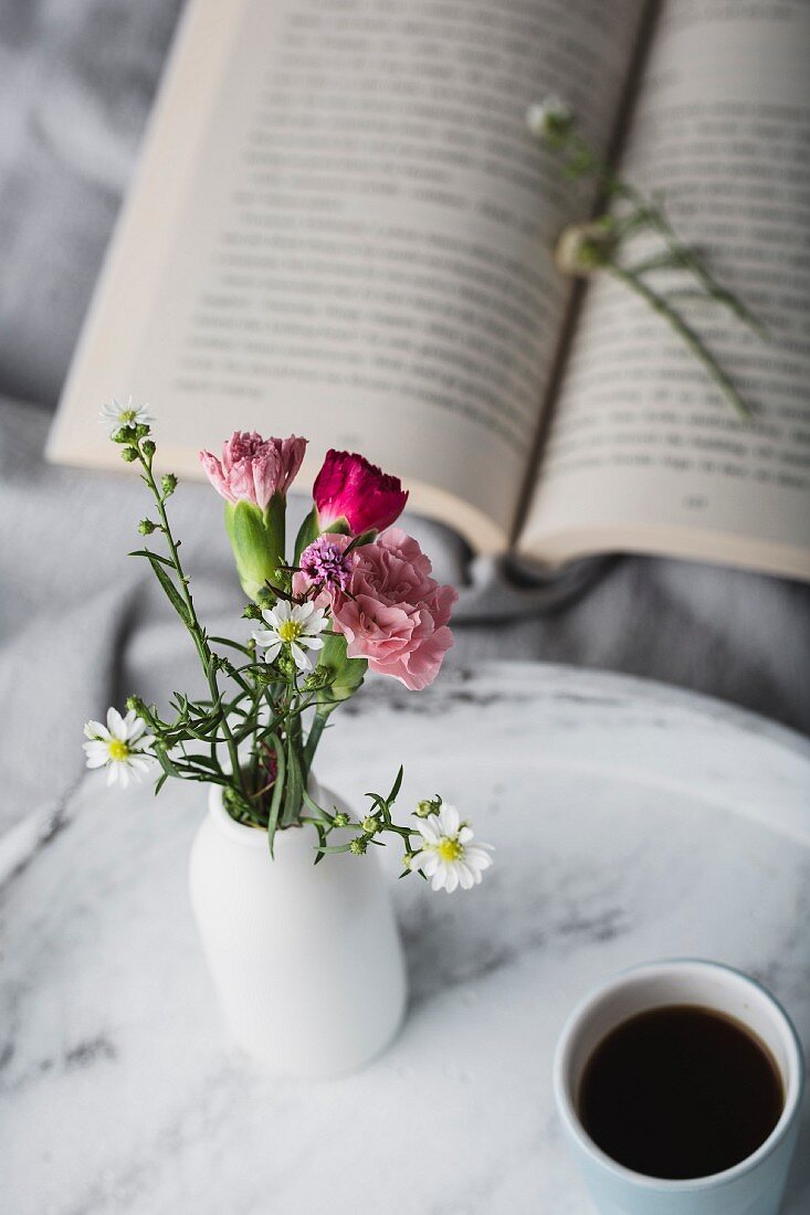 Open book, vase of flowers and cup of coffee on tray on bed
