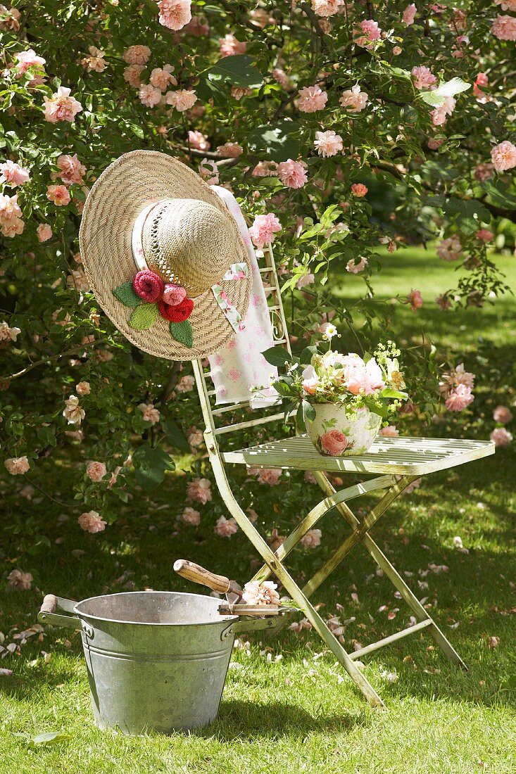 Vintage Gartenstuhl mit Blumengesteck und romantischem Strohhut neben Zinkeimer vor rosafarbener Kletterrose