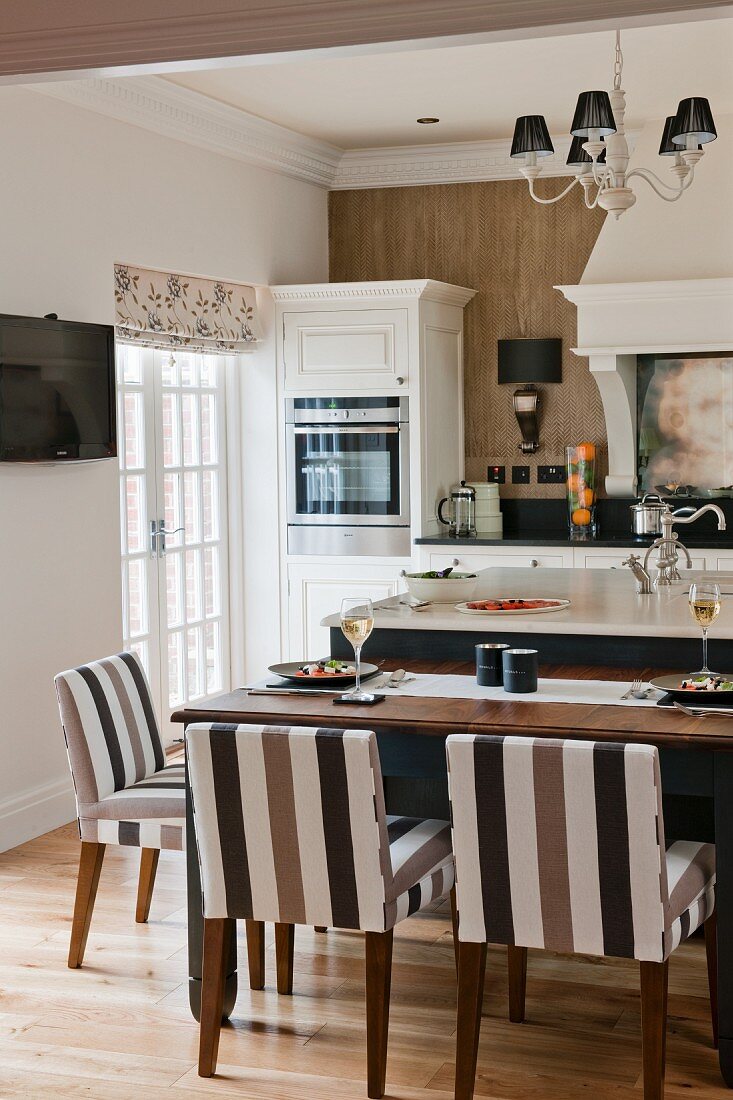 weiße Landhausküche mit Kücheninsel, gedecktem Esstisch mit braun gestreiften Polsterstühlen