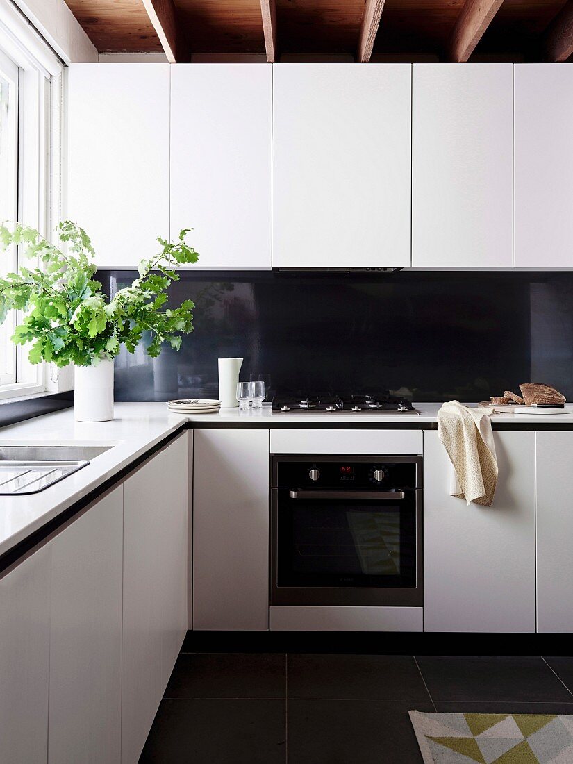 weiße Übereck-Küche mit schwarzem Spritzschutz und grünen Eichenblätterzweigen vor Fenster