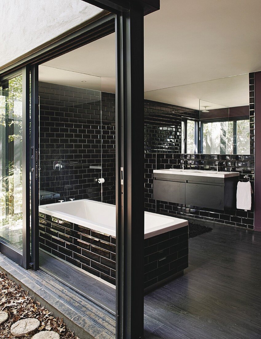 Bad mit schwarzen, glänzenden Wandfliesen, Badewanne und geöffneter Terrassenschiebetür