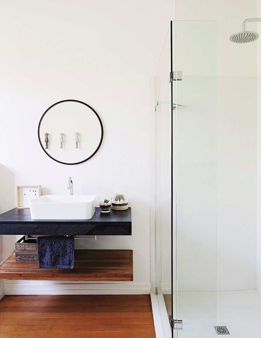 Verglaste Duschkabine neben rundem Wandspiegel und Waschbecken auf Waschtischplatte