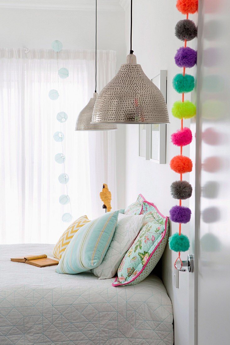 Colorful pompom garland on room door in bright bedroom with pendant lights and pastel-colored pillows
