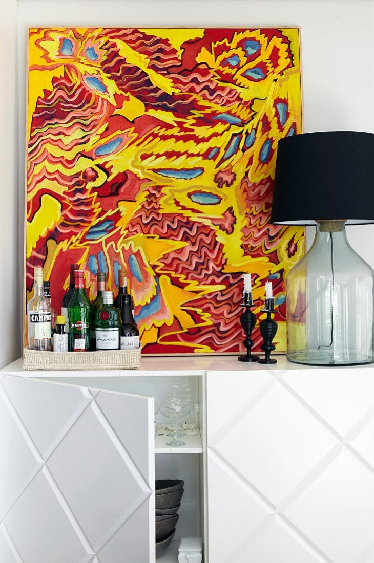 Colourful picture, bottles of spirits and table lamp on top of white sideboard