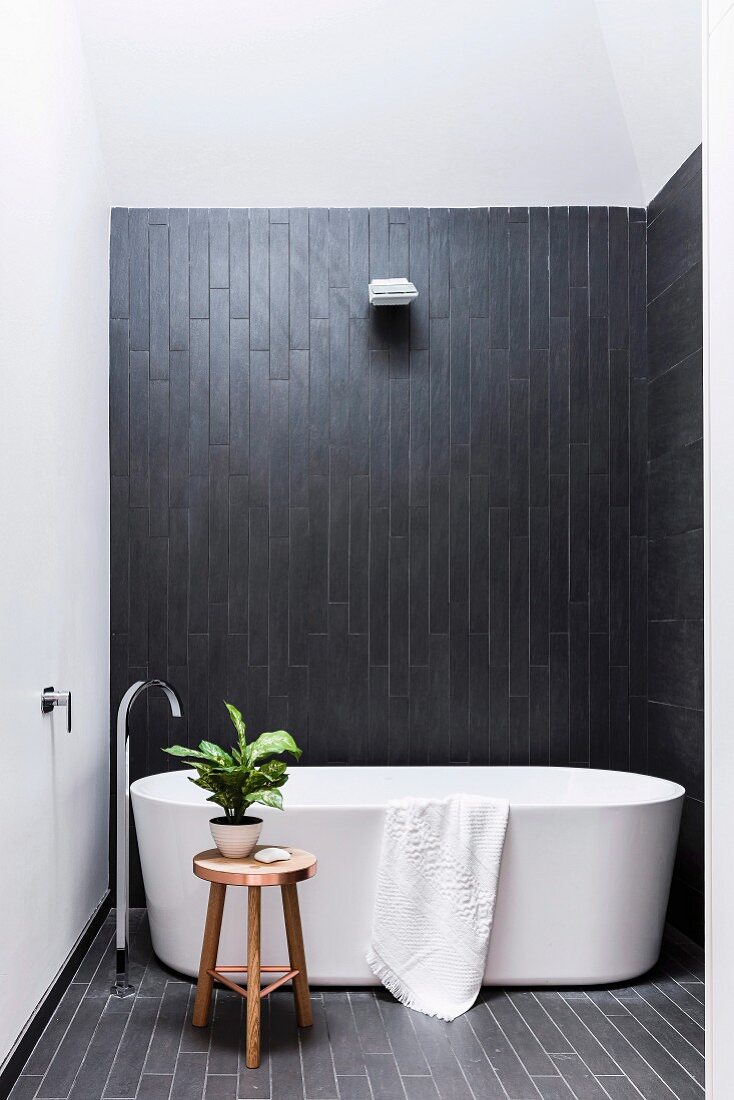 Freistehende Badewanne mit Standarmatur vor schwarzer Wandverkleidung in schlichtem Badezimmer