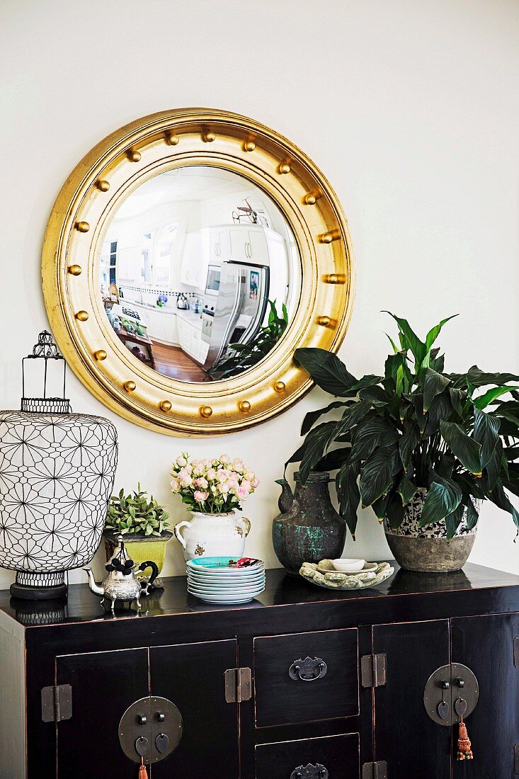 Round gold frame mirror above ethnic piece of furniture with lantern, green plant and stack of porcelain plates