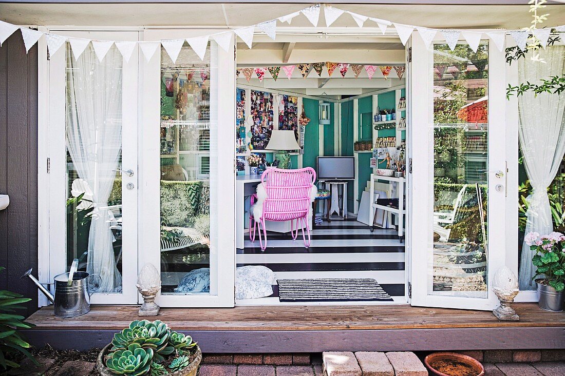Blick in eklektischen Wohnbereich mit rosafarbenem Korbsessel, auf Wimpelketten und geöffnete Fenstertüren