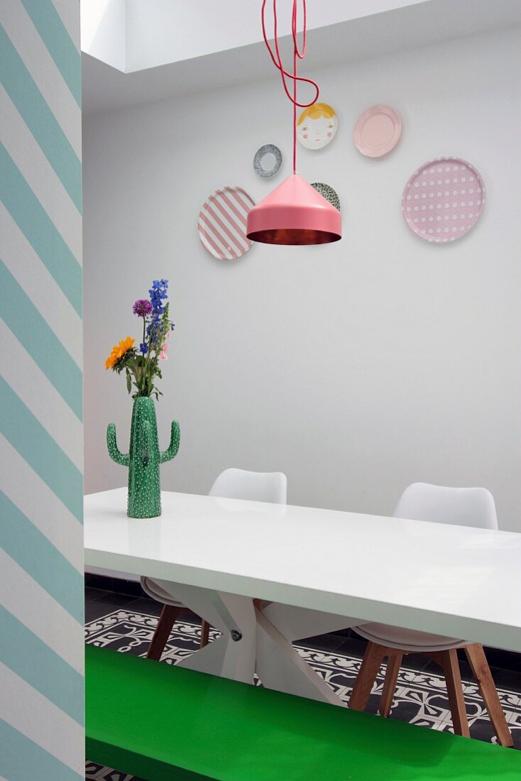 Weisser Esstisch mit Vase in Kaktusform vor weisser Wand mit pastellfarbenen Dekotellern