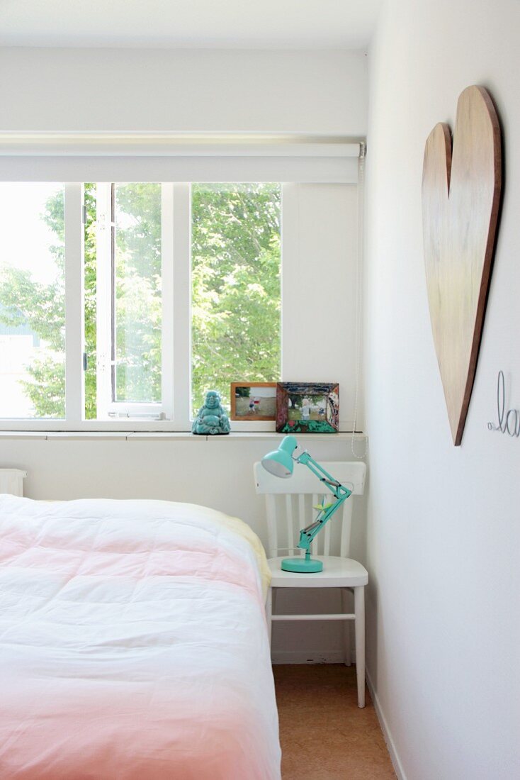 Türkisfarbene Retrolampe auf weißem Holzstuhl als Nachttisch neben Bett vor geöffnetem Fenster