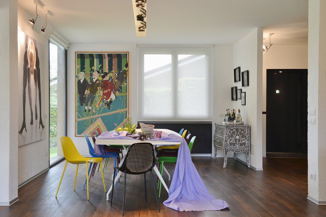 Colourful chairs, length of lilac fabric, artistic ethnic cabinet and modern artwork in dining area